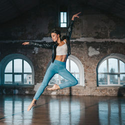 Young woman jumping in casual style - jeans and leather jacket doing ballet in old studio. 