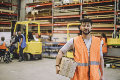 Young carpenter in reflective clothing carrying cardboard box in warehouse