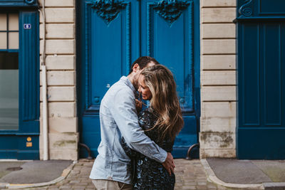 Side view of happy young couple in casual clothes hugging and kissing while standing against aged stone building with blue doors on city street