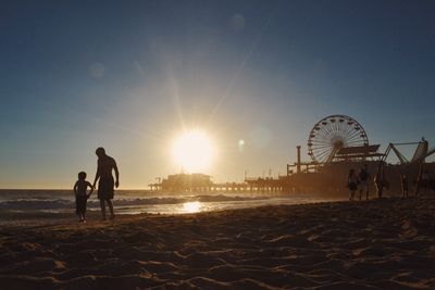 People walking at beach by santa monica pier against clear sky during sunset