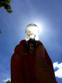 Cropped hand holding light bulb against sky in sunny day