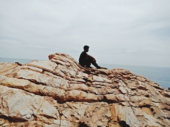 Side view of man sitting on rocks by sea against sky