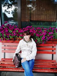 Full length of woman sitting on bench against red flowering plants