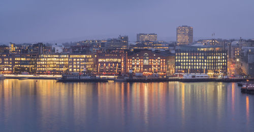 Illuminated buildings by river against sky in city at dusk