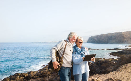 Smiling couple using digital tablet by sea against sky