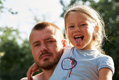 Portrait of daughter sticking out tongue with father against tree