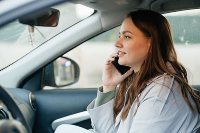 Beautiful girl with long hair in a grey trench coat using smartphone call gets into the car