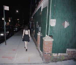 Rear view of woman walking on footpath amidst buildings in city alone at night