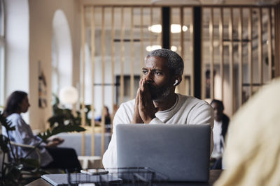 Worried businessman with hands clasped talking through wireless in-ear headphones at office