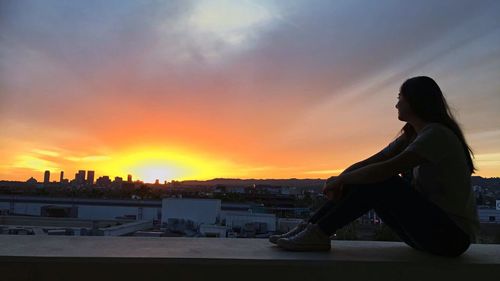Woman sitting in city against sky during sunset