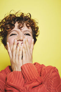 Amazed laughing female with curly hair wearing orange knitted sweater covering mouth with hands and looking away while standing against yellow background