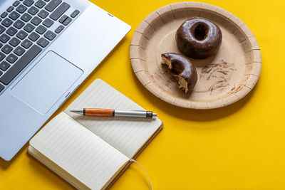 Bitten chocolate donuts in a cardboard plate, next to a laptop on yellow bacground