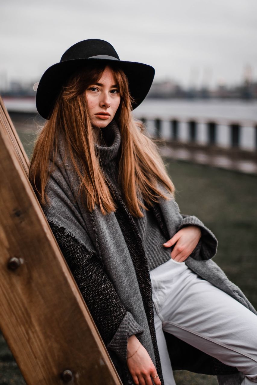 hat, clothing, real people, one person, hair, leisure activity, lifestyles, long hair, hairstyle, young adult, focus on foreground, water, young women, day, standing, three quarter length, beautiful woman, women, looking away, outdoors, warm clothing