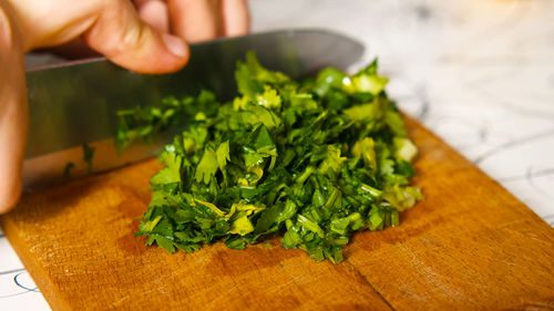 A close-up of a woman's hand with a knife is chopping parsley on a cutting board.