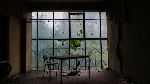 Table and empty chair in abandoned office in front of big window with broken glass