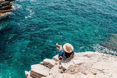 Rear view of young woman wearing summer style clothes and hat, sitting on edge of cliff above sea.