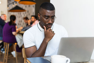 Thoughtful black adult male in eyeglasses and casual clothes sitting in chair at table in light cafeteria and browsing on netbook near cup of coffee with people on background