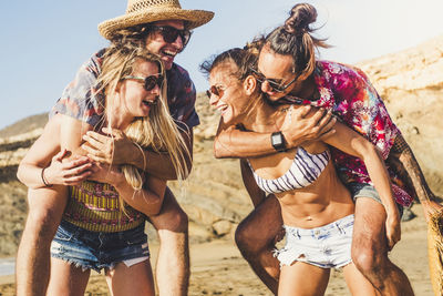 Girlfriends piggybacking boyfriends at beach against sky