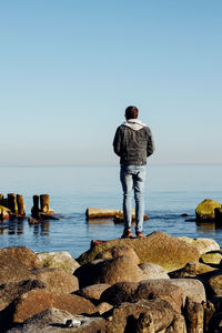 Full length rear view of man standing on rocks by sea against clear sky