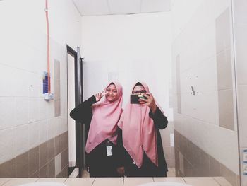 Portrait of smiling woman gesturing while friend taking selfie while reflecting on mirror