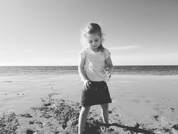 Full length of cute girl playing with sand while standing at beach against sky during sunny day