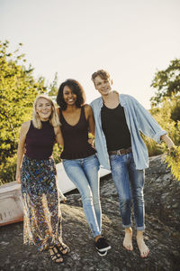 Full length portrait of young friends standing on rock at harbor against sky