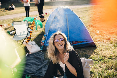 High angle view of cheerful woman wearing sunglasses sitting against tent on field during summer