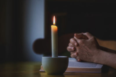 Close-up of hand praying over book by illuminated candle on table