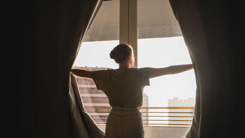 Back view of unrecognizable young female in casual clothes opening blackout curtains and admiring modern city during holidays in hotel