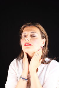 Close-up of woman with eyes closed touching neck against black background