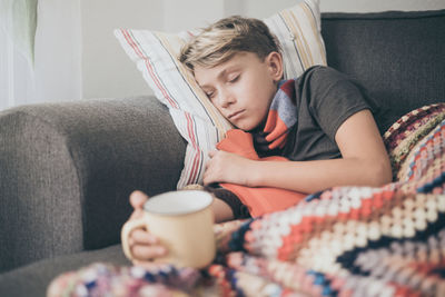 Ill boy having drink in cup while lying on sofa at home