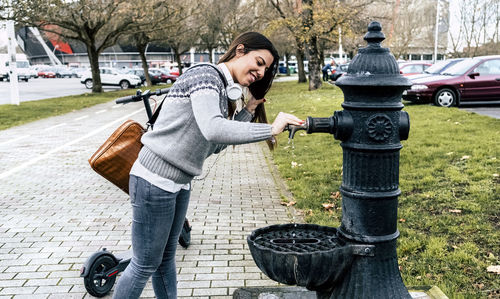 Smiling young woman with electric scooter drinking water from a well in the city