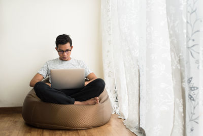 Young man using laptop while sitting on bean bag by window at home