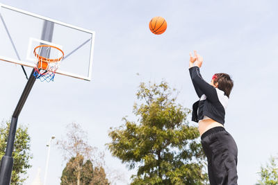 Low angle side view of female player throwing ball into hoop while playing basketball against cloudy sky and green trees