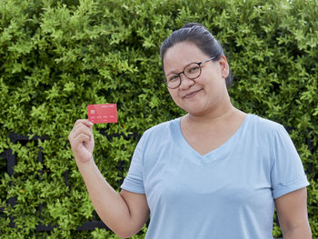 Portrait of smiling woman holding credit card while standing against plants