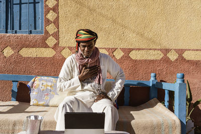 Sincere egyptian male in traditional turban and robe having video chat on tablet while sitting on sofa in backyard on sunny day