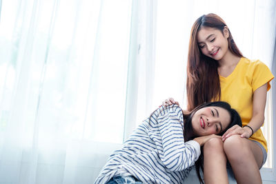 Smiling woman lying on friend laps against curtain at home