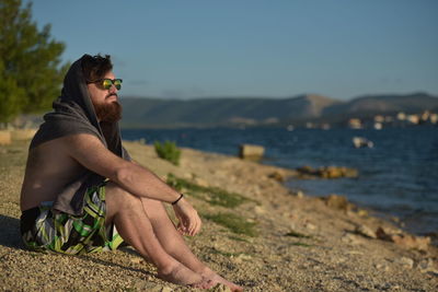Bearded man sitting at beach against sky
