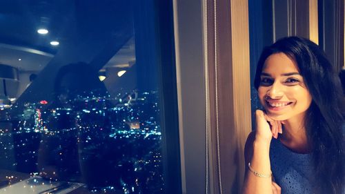 Portrait of smiling woman by window against illuminated cityscape at night