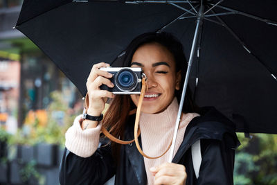 Smiling woman carrying umbrella photographing through camera in city