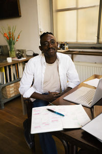 Portrait of male entrepreneur sitting by table with laptop and diary in workplace