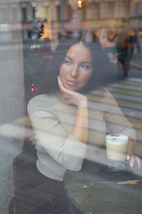 Thoughtful woman with coffee sitting in restaurant seen through window