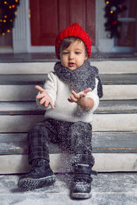 Child in a white sweater and a red knitted hat sits on the steps with snow at home at christmas