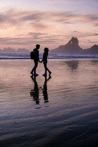 Rear view of man walking at beach against sky during sunset