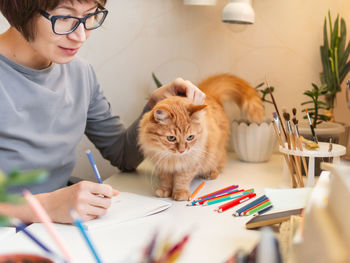 Woman with short hair cut is drawing in notebook. cute ginger cat sits near her. pet and artist.