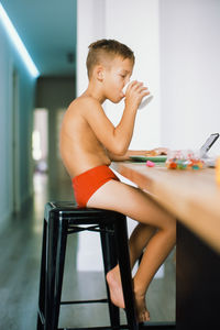 Side view of shirtless boy drinking drink while using digital tablet at home