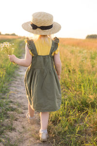 A little blonde girl in a straw hat walks in a field with a bouquet of daisies. 