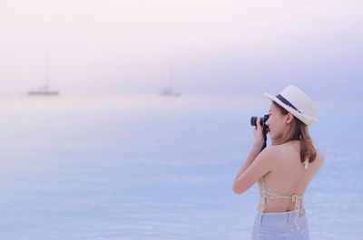 Woman photographing while standing at beach against sky during sunset