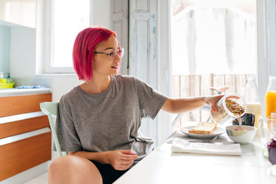 Optimistic young woman with dyed hair smiling and enjoying healthy dish while sitting at table in cozy kitchen at home in morning