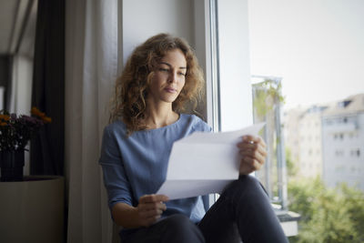 Woman reading paper while sitting on window sill at home